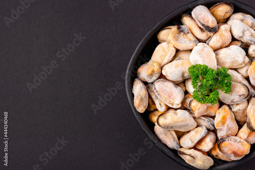 Frozen mussels meat with green parsley in a bowl on a black background, close-up top view.