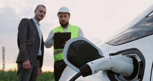 Young man talks to engineer showing tablet near green field focus on contemporary electromobile with plugged charger closeup photo