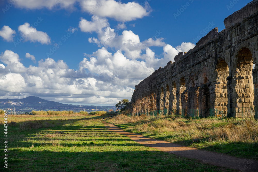 Naklejka premium Parco degli Acquedotti, Rome.The imposing arches of the archaeological area the water system of ancient Rome.Park of the Via Appia, the Castelli Romani in the distance with blue sky, clouds and shadow