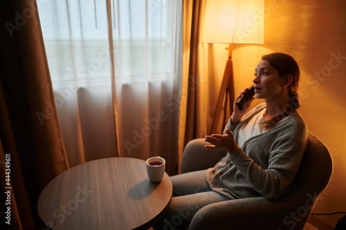 Communication. A woman is sitting in a chair in the living room and talking on the phone. There is a cup of coffee on the table in front of her. There is a floor lamp in the corner of the room.