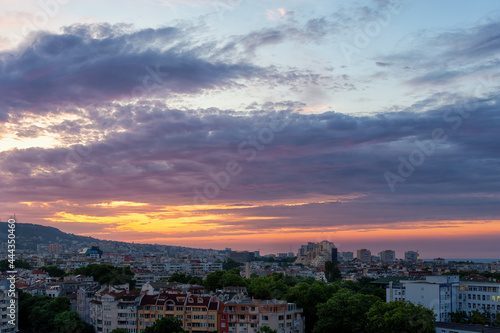Orange dawn sky with purple blue picturesque clouds over the sleeping city. Pastel colored sunrise heaven over Varna. Calm cloudy dawn flares up in the sky. Early morning summer cityscape. © Maryia