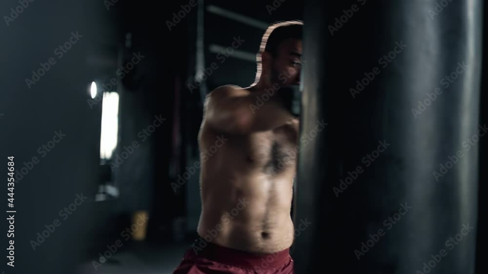 A muscular alone boxer is hits punching bag in the gym. Sportsman is boxing in dark hall. Self confident man is training indoors. Maximum focus and strength. Concept of lifestyle health and sports. A muscular alone boxer is hits punching bag in the gym. Sportsman is boxing in dark hall. Self confident man is training indoors. Maximum focus and strength. Concept of lifestyle health and sports.