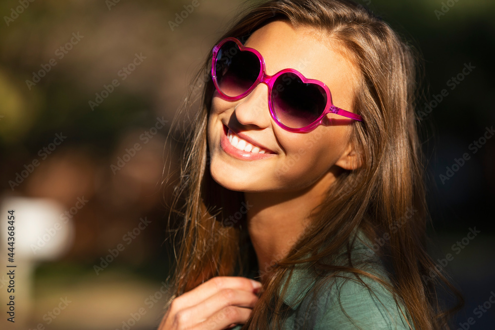 Portrait of young woman standing outside in bright sun wearing heart shaped sunglasses, looking into camera lens Portrait of young woman standing outside in bright sun wearing heart shaped sunglasses, looking into camera lens