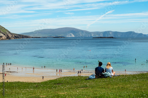 Beautiful couple sitting on a grass and enjoy great view on the ocean. Keem bay and beach, Achill island, Ireland. Warm sunny day. Love and romance concept. Travel and outdoor activity