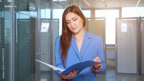 Concentrated brunette businesswoman looks through company report sheets of papers in blue folder standing in office hall against windows, sunlight