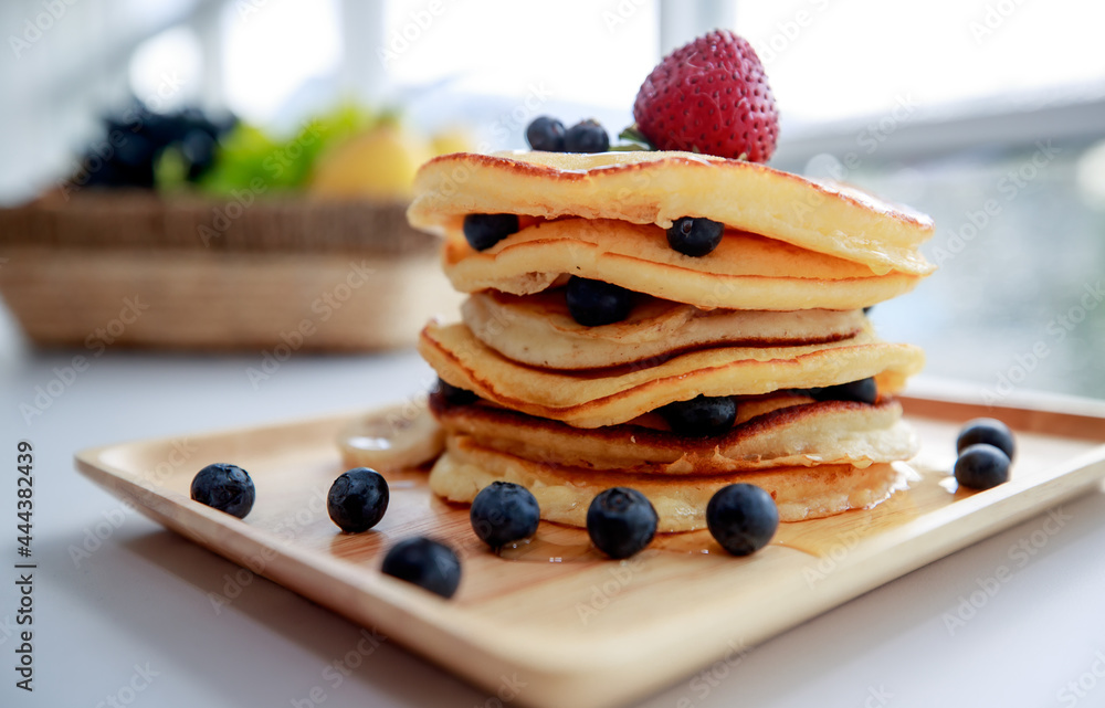 in kitchen on bright morning, pancake tower on wooden plate topping by strawberry and blueberry look very sweet tasty prepared to be served as superb healthy dessert after breakfast