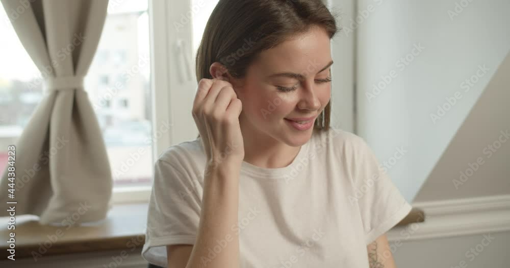 A cute brunette girl of Slavic appearance sits in a white T-shirt at the desk, on which there is a laptop, takes an earpiece, sticking it in her ear, listening to music in headphones and dancing A cute brunette girl of Slavic appearance sits in a white T-shirt at the desk, on which there is a laptop, takes an earpiece, sticking it in her ear, listening to music in headphones and dancing