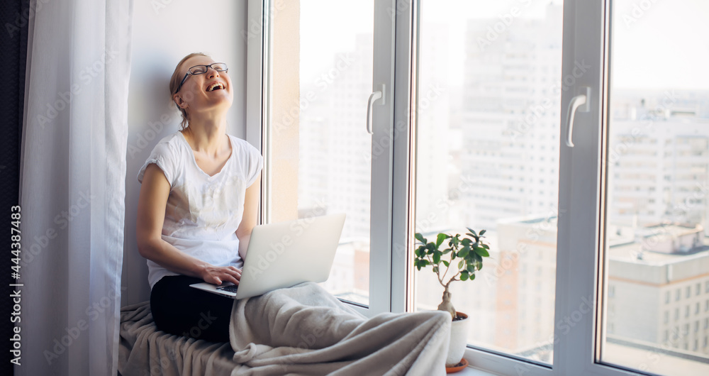 Happy young adult woman freelancer wearing glasses sitting on window background. Laughing blonde working on laptop. Workplace on the windowsill. Home office concept. Happy young adult woman freelancer wearing glasses sitting on window background. Laughing blonde working on laptop. Workplace on the windowsill. Home office concept.