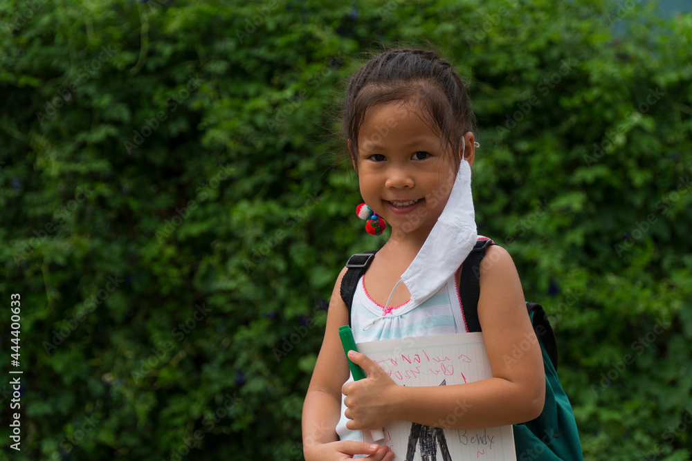 Little asian girl wearing face mask with green back pack holding stationery in green nature park background. Back to school concept. Little asian girl wearing face mask with green back pack holding stationery in green nature park background. Back to school concept.