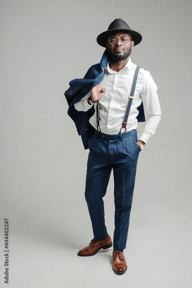 Full-length photo. Attractive stylish african american businessman in a hat and with suspenders holds a jacket with his hand on his shoulder Full-length photo. Attractive stylish african american businessman in a hat and with suspenders holds a jacket with his hand on his shoulder