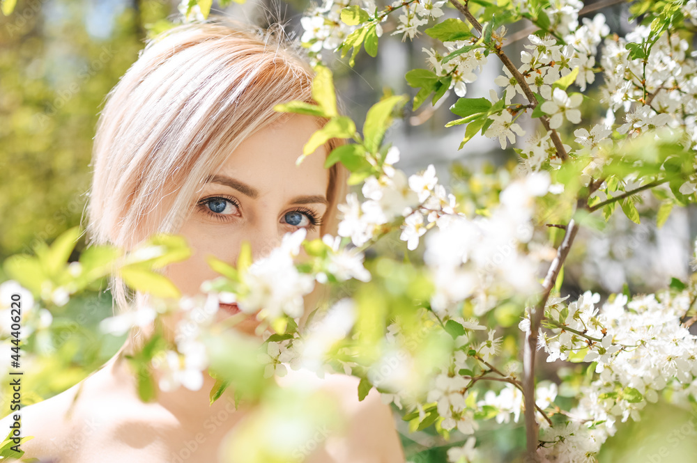 Fototapeta premium Close up young beautiful blonde woman with blue eyes posing in blooming spring garden with white flowers. Blooming apple or cherry tree. Concept of natural cosmetics, youth, healthy skin and teeth