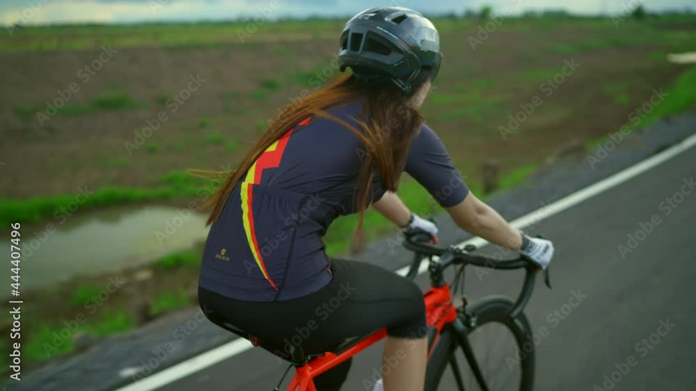 Back view tracking.Asian women Orange cyclist wearing protective helmet exercise training fast riding a bike on the roads outside the city Back view tracking.Asian women Orange cyclist wearing protective helmet exercise training fast riding a bike on the roads outside the city
