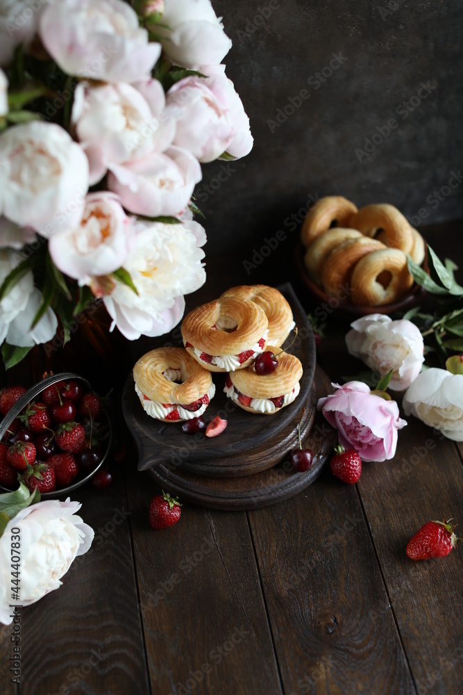 Custard rings with curd cream and berries on a dark table with a bouquet of peonies. Still life with flowers and sweets. Custard rings with curd cream and berries on a dark table with a bouquet of peonies. Still life with flowers and sweets.