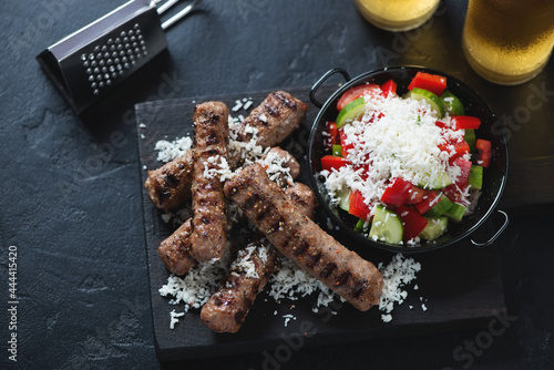 Grilled cevapi or serbian skinless beef sausages with grated bryndza and shopska salad, elevated view on a black stone background, horizontal shot photo
