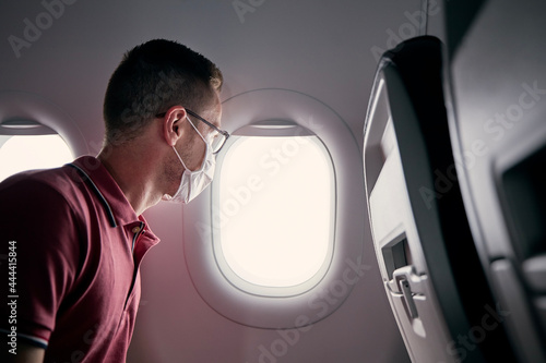 Passenger with protective face mask in airplane looking through window. Themes traveling in new normal and personal protection during pandemic covid-19..