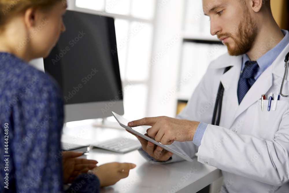Unknown bearded doctor and patient woman discussing current health examination while sitting and using tablet computer, close-up. Medicine concept Unknown bearded doctor and patient woman discussing current health examination while sitting and using tablet computer, close-up. Medicine concept