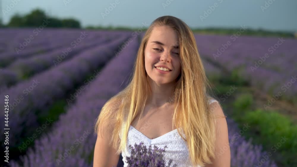 Young smiling caucasian woman holding a bouquet of fragrant flowers looking at the camera, lavender field on the background. Happy woman takes a deep breath, sniffs flowers outdoors. Young smiling caucasian woman holding a bouquet of fragrant flowers looking at the camera, lavender field on the background. Happy woman takes a deep breath, sniffs flowers outdoors.