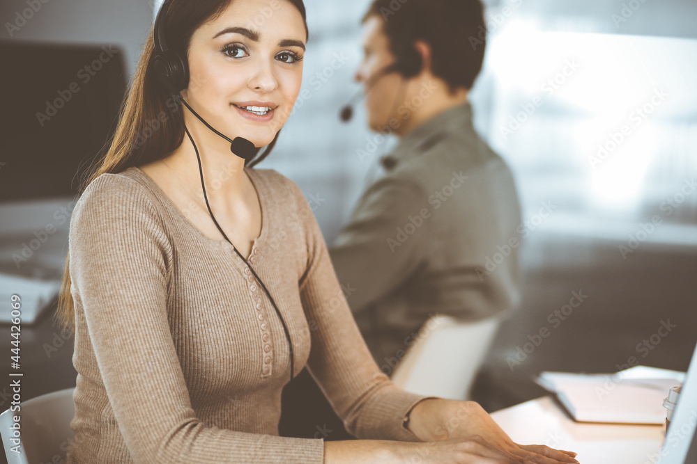 Fototapeta premium Young friendly girl in headsets is talking to a firm's client, while sitting at the desk in a modern office together with her colleague. Call center operators at work