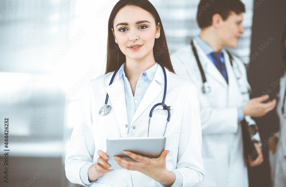 Smiling intelligent woman-doctor is holding a tablet computer in her hands, while she is standing together with her colleagues in a clinic. Physicians at work. Perfect medical service in a hospital Smiling intelligent woman-doctor is holding a tablet computer in her hands, while she is standing together with her colleagues in a clinic. Physicians at work. Perfect medical service in a hospital
