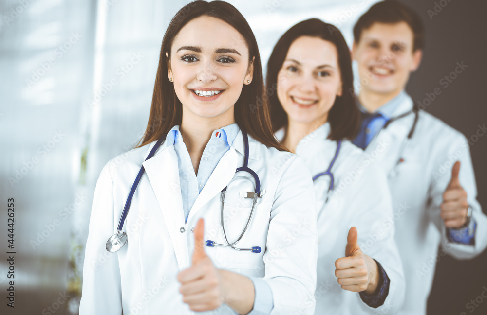 Group of young cheerful doctors is standing as a team with thumbs up in a hospital office and is ready to help patients. Medical help, insurance in health care, best disease treatment and medicine Group of young cheerful doctors is standing as a team with thumbs up in a hospital office and is ready to help patients. Medical help, insurance in health care, best disease treatment and medicine