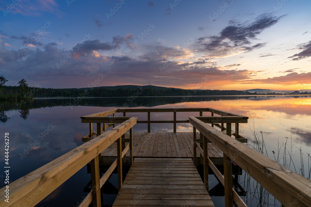wooden pier leads out onto a calm lake with forest on the opposite shore and a colorful sunset sky