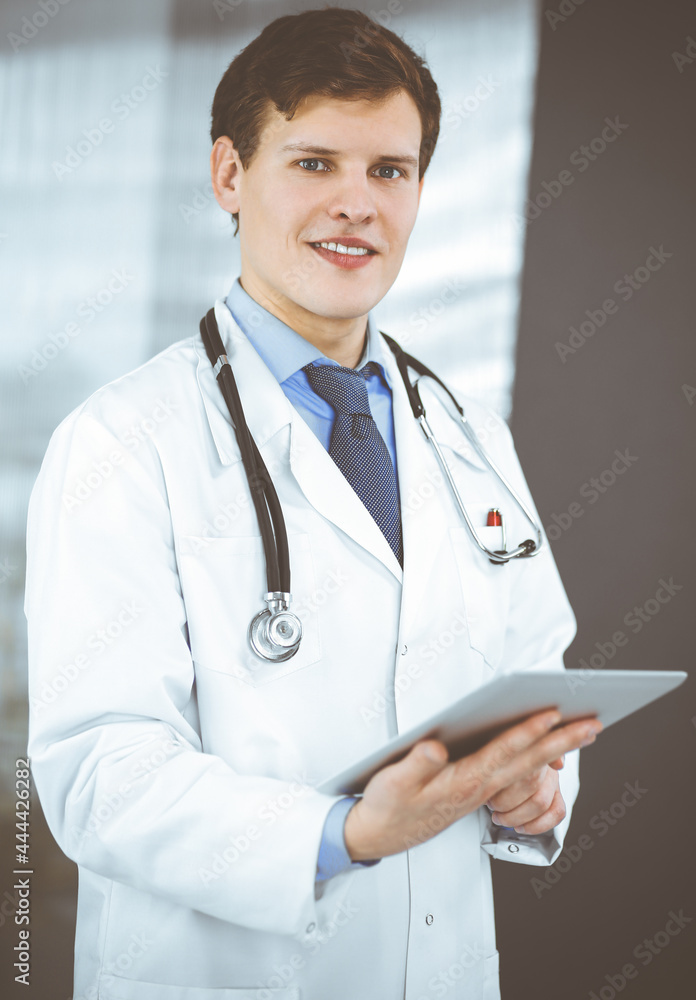 Young friendly doctor is checking some information on his computer tablet. Portrait of a professional physician at work in a clinic. Medicine concept Young friendly doctor is checking some information on his computer tablet. Portrait of a professional physician at work in a clinic. Medicine concept