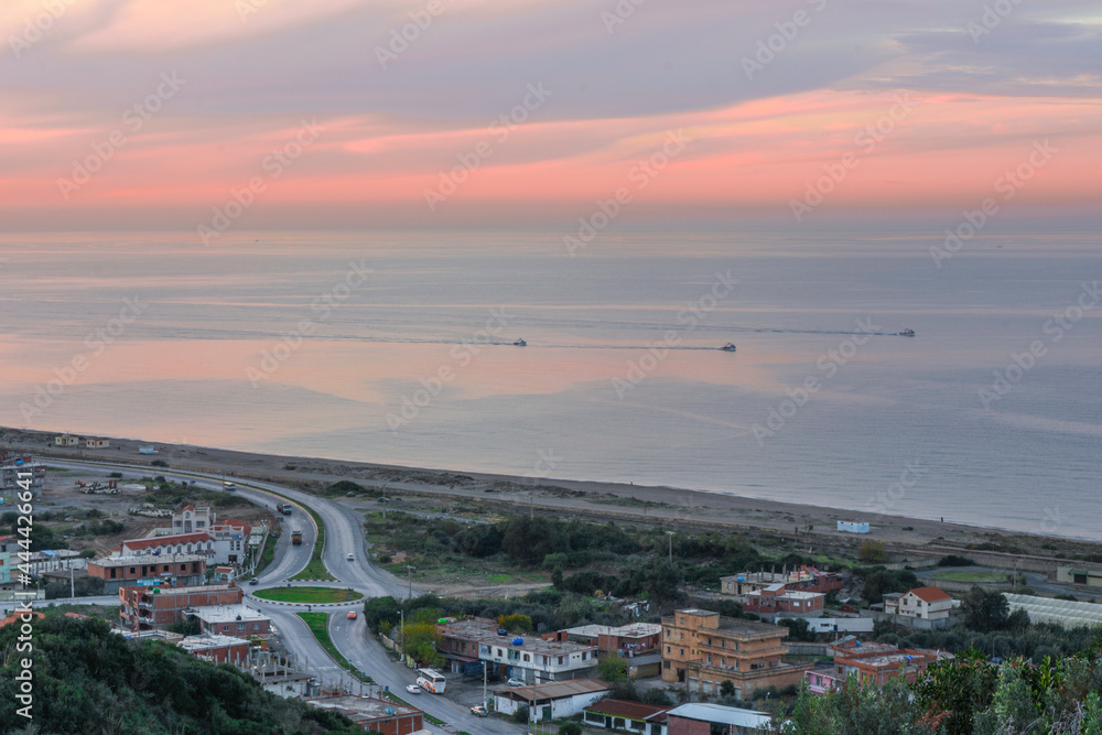 Aerial view of a highway near the beach in the Mediterranean Sea, a roundabout road near the coast and three fishing boats in the sea at sunset, a coastal village in Jijel Province, Algeria, Africa.