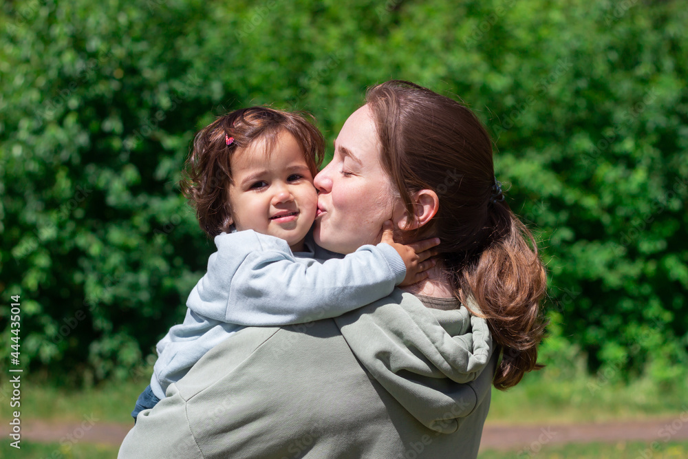 A young caucasian woman kisses the child on the cheek while holding her in arms. A young mother walks with her daughter in a park in nature. A little girl hugs her mom tightly. Family, love. Sunny day A young caucasian woman kisses the child on the cheek while holding her in arms. A young mother walks with her daughter in a park in nature. A little girl hugs her mom tightly. Family, love. Sunny day