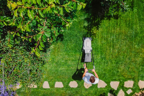 Caucasian man pushing lawn mower for cutting green grass in garden with sunlight at summer season. Aerial view. Housework and lifestyle concept. photo