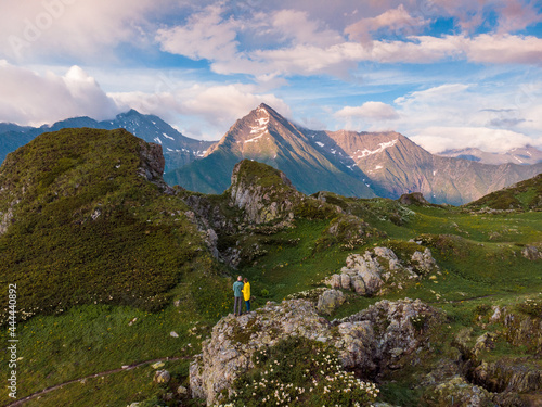A couple stands on among the flowering mountain meadows and enjoys the views of the mountain peaks while filming themselves on a drone. Aerial view of the Caucasus Mountains in Krasnaya Polyana