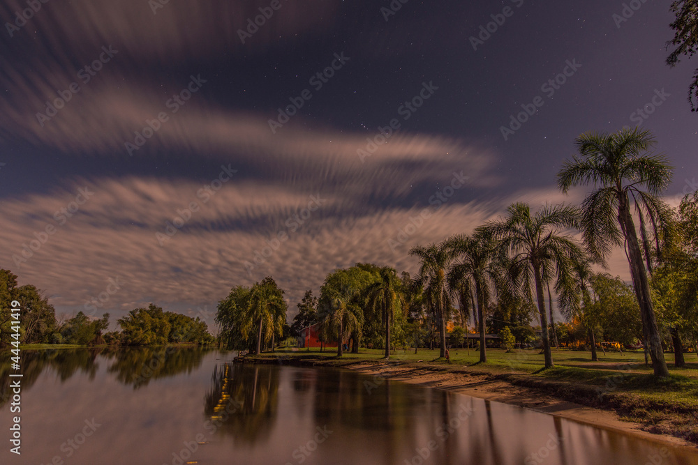 Night shot of a quiet riverside and palm trees in the shore with moving clouds. Stars can be seen in the sky and some clouds in the horizon. Villa Paranacito, Entre Ríos, Argentina