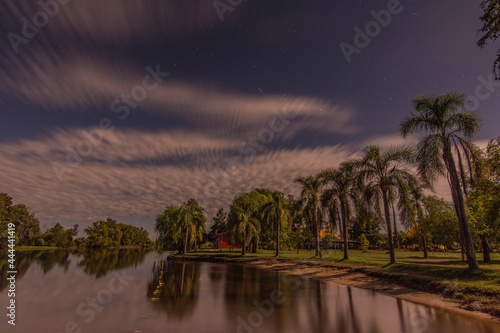 Night shot of a quiet riverside and palm trees in the shore with moving clouds. Stars can be seen in the sky and some clouds in the horizon. Villa Paranacito, Entre Ríos, Argentina