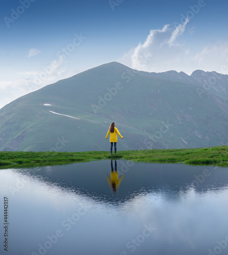 A traveller girl in a yellow jacket stands on the shore of a lake. Beautiful reflection of a woman in the water of a mountain lake photo
