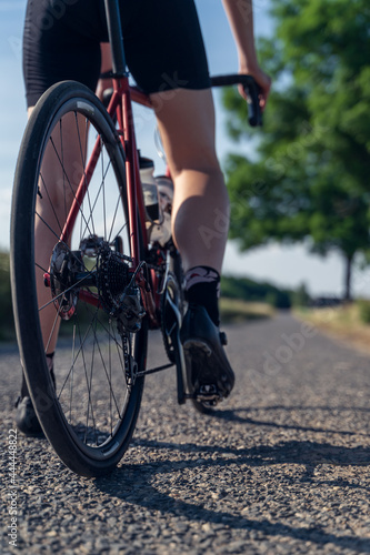 Vertical close up of a rear wheel, and the feet of a woman cyclist, standing still, ready to start her bike ride. In the background a paved road in the middle of a field.