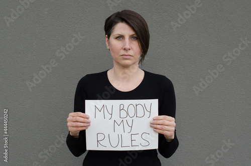 Tela Middle age woman with short black hair looking at camera, holding banner My Body My Rules in front of her
