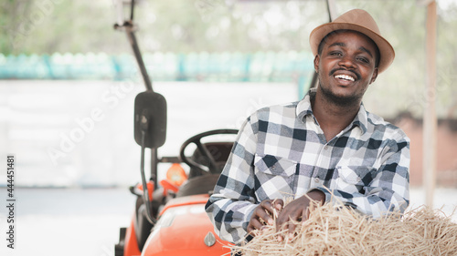 African farmer standing and smiling with rice straw bales and tractor. Agriculture or cultivation concept