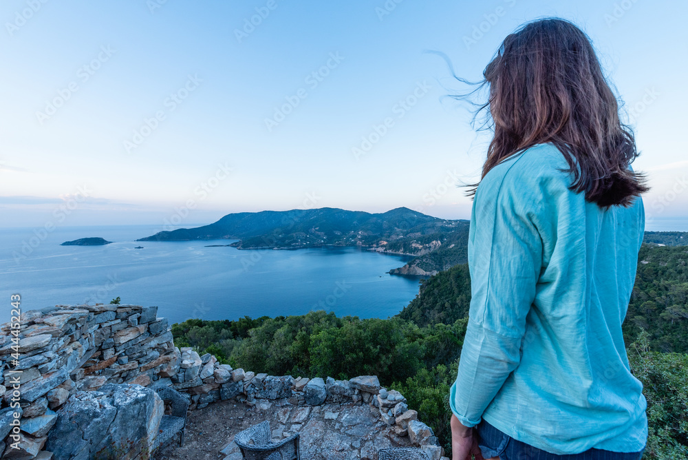 Woman on her back standing over the coastline landscape at dawn, with a blue sea, green vegetation and a clear sky Woman on her back standing over the coastline landscape at dawn, with a blue sea, green vegetation and a clear sky