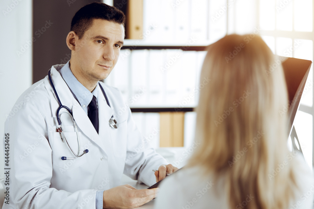 Friendly male doctor and patient woman discussing current health examination while sitting in sunny clinic. Perfect medical service in hospital Friendly male doctor and patient woman discussing current health examination while sitting in sunny clinic. Perfect medical service in hospital