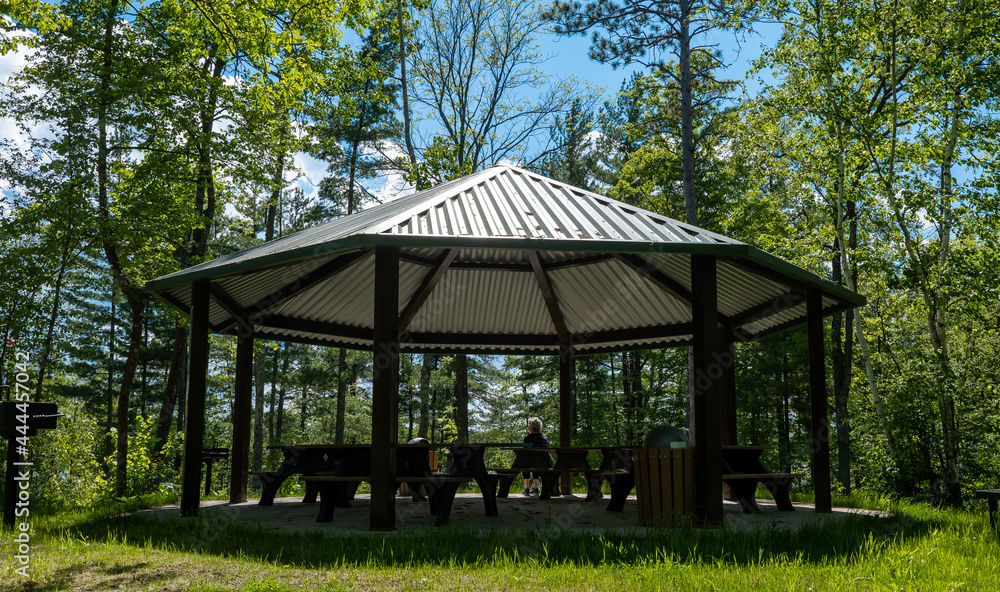 Gazebo with steel roof and picnic tables and surounded by trees on a mostly sunny day with blue sky and light fluffy clouds.