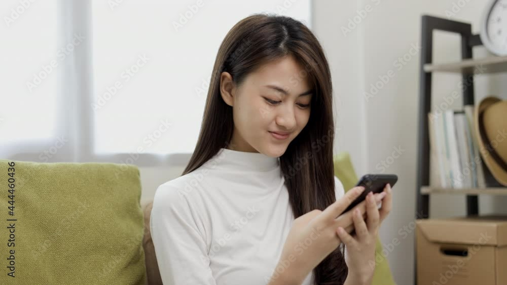 Happy young asian woman looking at screen of smartphone sitting on sofa in living room she shopping online. She very happy. Teenage girl reading news from mobile phone feeling excited. Happy young asian woman looking at screen of smartphone sitting on sofa in living room she shopping online. She very happy. Teenage girl reading news from mobile phone feeling excited.