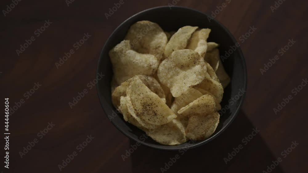 Slow motion handheld shot of organic potato chips with black pepper in black bowl on walnut table Slow motion handheld shot of organic potato chips with black pepper in black bowl on walnut table