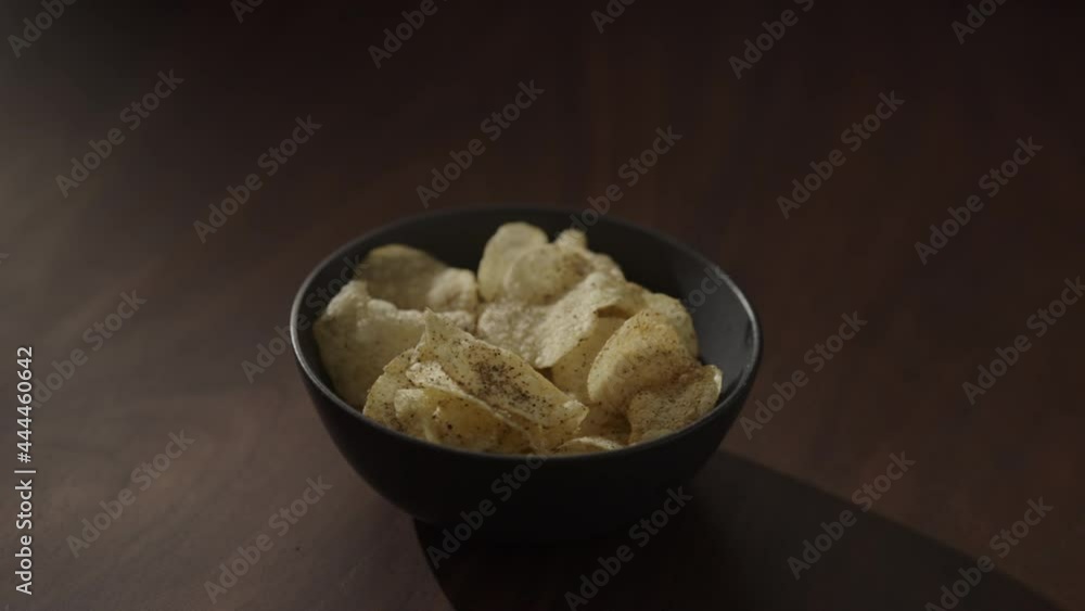 Slow motion handheld shot of organic potato chips with black pepper in black bowl on walnut table Slow motion handheld shot of organic potato chips with black pepper in black bowl on walnut table