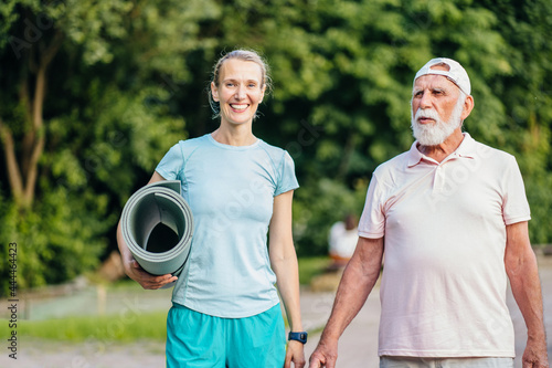 Active senior man and woman physiotherapist carrying exercise mats after sports workout in nature. Old man and his adult daughter holding yoga mats, walking along park path together, talking.