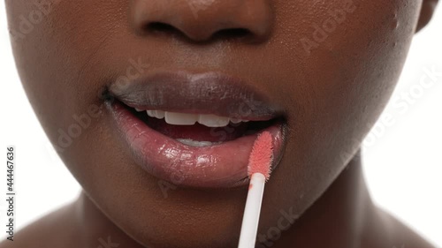 Close up of an african woman's mouth, applying lip gloss on her top and bottom lips. White background.
