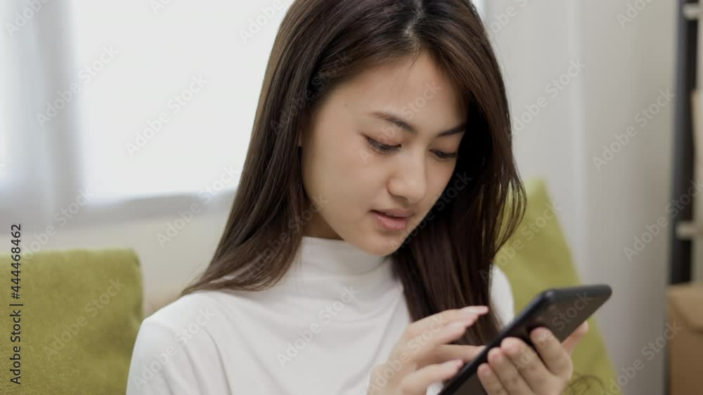 Happy young asian woman looking at screen of smartphone sitting on sofa in living room she shopping online. She very happy. Teenage girl reading news from mobile phone feeling excited. Happy young asian woman looking at screen of smartphone sitting on sofa in living room she shopping online. She very happy. Teenage girl reading news from mobile phone feeling excited.