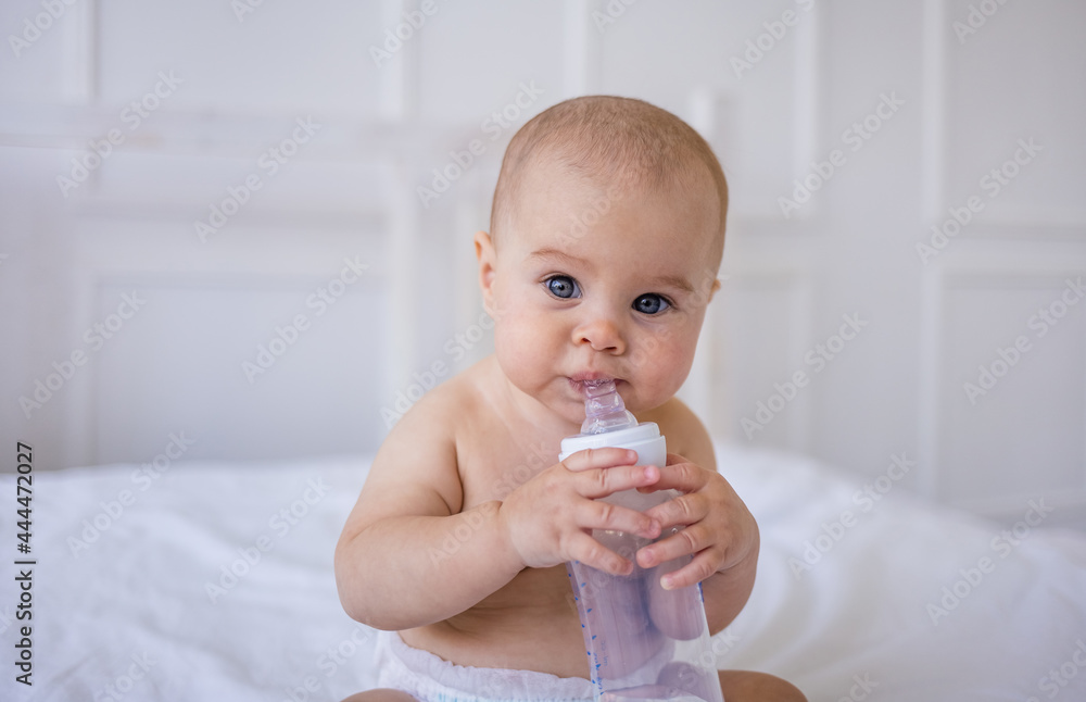 portrait of a baby girl sitting and drinking a water bottle on a white bed in a room. Child care portrait of a baby girl sitting and drinking a water bottle on a white bed in a room. Child care