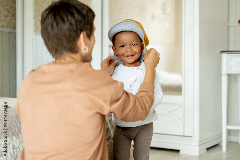 Loving mommy showing cute dino hat to her sweet afro-american son, friendly family preparing going for a walk, choosing clothes together, upbringing psychology Loving mommy showing cute dino hat to her sweet afro-american son, friendly family preparing going for a walk, choosing clothes together, upbringing psychology