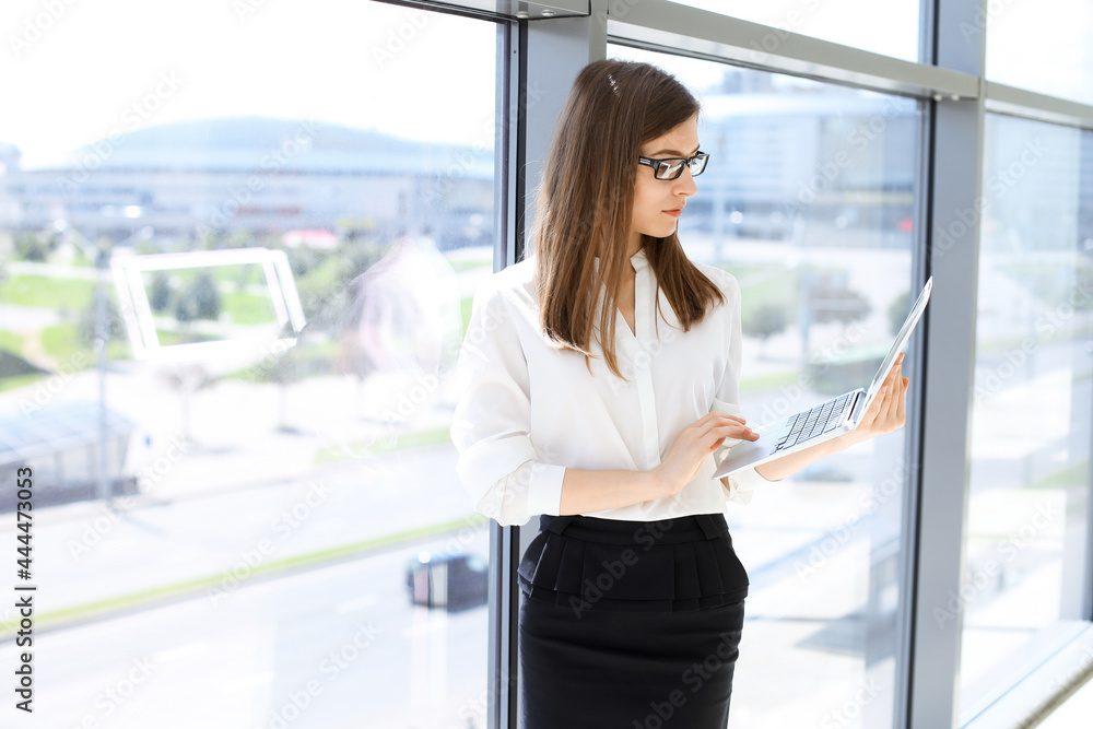 Beautiful female specialist with laptop computer standing in modern office and smiling charmingly. Working on design, data analysis, plan strategy. Business people concept Beautiful female specialist with laptop computer standing in modern office and smiling charmingly. Working on design, data analysis, plan strategy. Business people concept