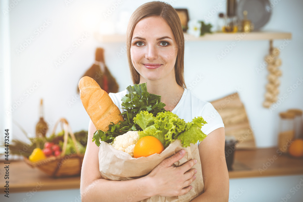 An attractive young woman holding the paper bag full of vegetables while standing and smiling in sunny kitchen. Cooking concept An attractive young woman holding the paper bag full of vegetables while standing and smiling in sunny kitchen. Cooking concept