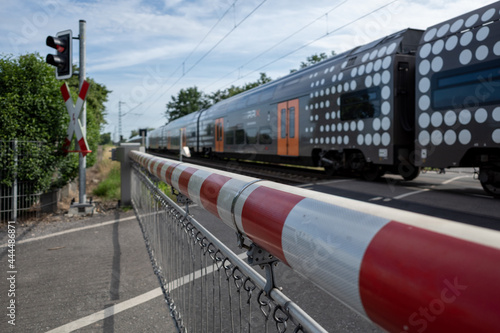 Fotografi Selective focus view at red and white level crossing railway barrier which block the road and regional train move on the rail on countryside in Germany